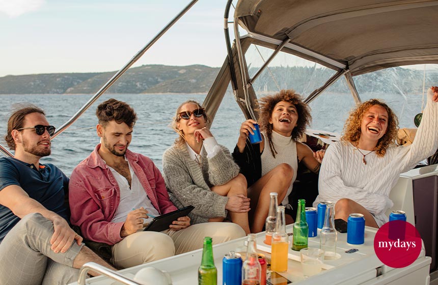 Eine Gruppe von Freunden sitzen lachend mit Getränken unter einem Dach auf einem Segelboot 