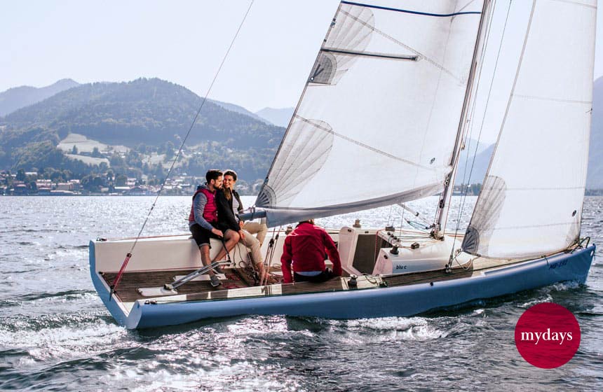 Ein Segelboot auf einem See mit Bergen im Hintergrund. Ein zufriedenes Paar und eine 3. Person segeln dem Horizont entgegen