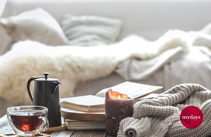 Ein Buch liegt auf einem Couchtisch mit einer Tasse Tee und einer Wolldecke. Im Hintergrund befindet sich eine Couch.